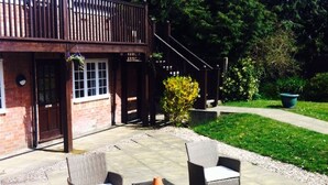 A patio with wicker furniture and a small table, a brick building with a wooden balcony, and a well-maintained lawn. - The Charlton Inn (Blandford Forum)