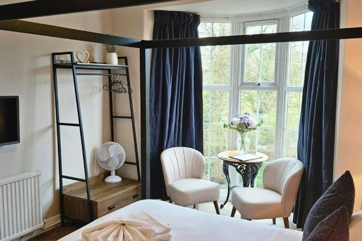 Ein Bett mit Baldachin, zwei Sessel, ein kleiner Tisch mit einer Vase und ein Ventilator in einem Zimmer mit einem großen Fenster und Vorhängen.