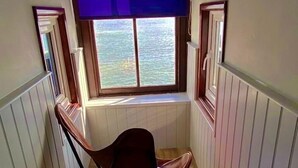 Una habitación estrecha con ventana que da a una masa de agua, una silla plegable y paneles de madera en las paredes.