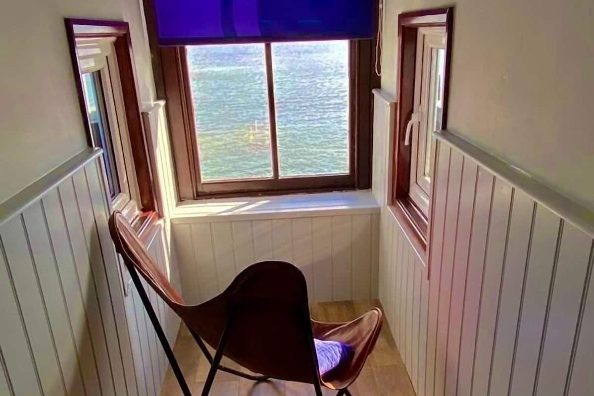 Una habitación estrecha con ventana que da a una masa de agua, una silla plegable y paneles de madera en las paredes.