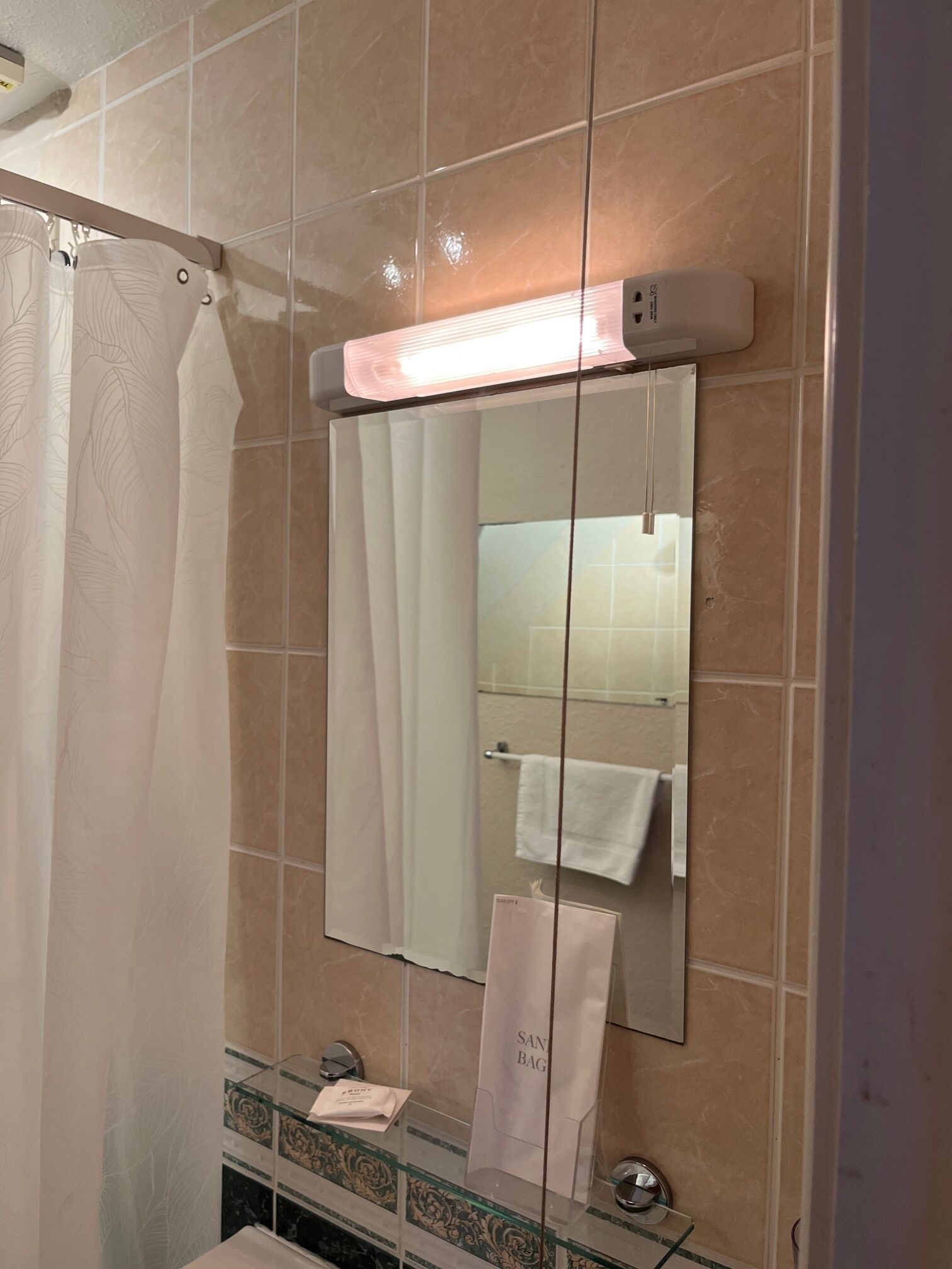 A bathroom with a shower curtain, a towel rack with a white towel, a glass shelf with toiletries, and a mirror with a light above it.