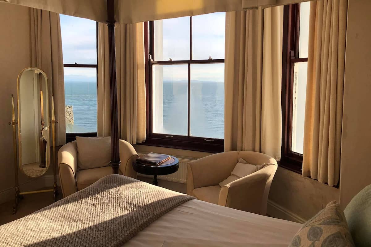 Habitación de hotel con cama, dos sillones, una mesa redonda y un ventanal con vistas al mar.