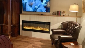 Una sala de estar con chimenea, un sillón de cuero, un televisor sobre la chimenea y una lámpara en una mesita auxiliar.