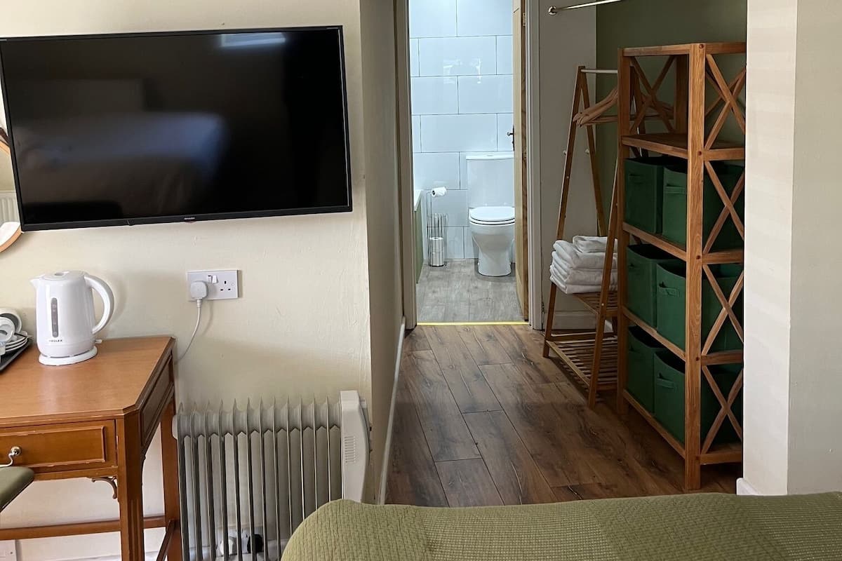 Una habitación con televisión, un radiador, una mesa de madera con hervidor, una cama verde y un estante de madera con cajas de almacenamiento.