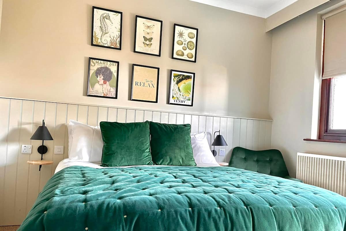 Un dormitorio ordenado con una cama grande, ropa de cama verde y una serie de láminas botánicas enmarcadas en la pared.