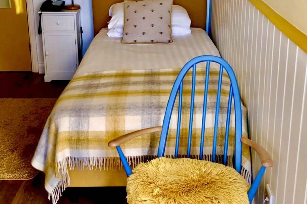 Un dormitorio con cama, una silla azul, una lámpara, una mesita pequeña y un radiador.