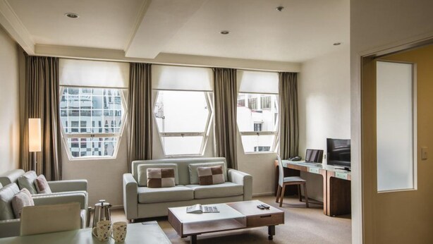 A modern hotel room with a sofa, a coffee table, a desk with a computer, and large windows offering a city view. - Quest on Lambton (Wellington)