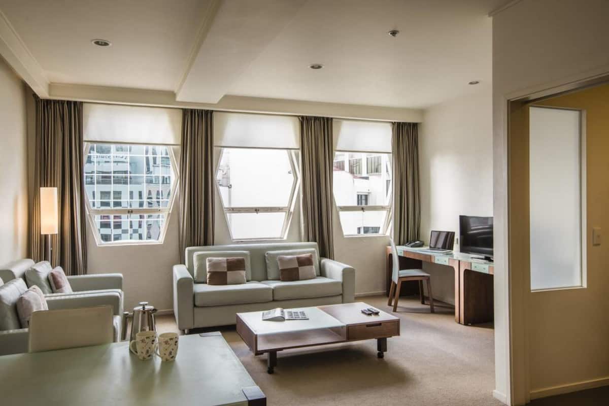 Une chambre d’hôtel moderne équipée d’un canapé, d’une table basse, d’un bureau avec un ordinateur et de grandes fenêtres donnant sur la ville.