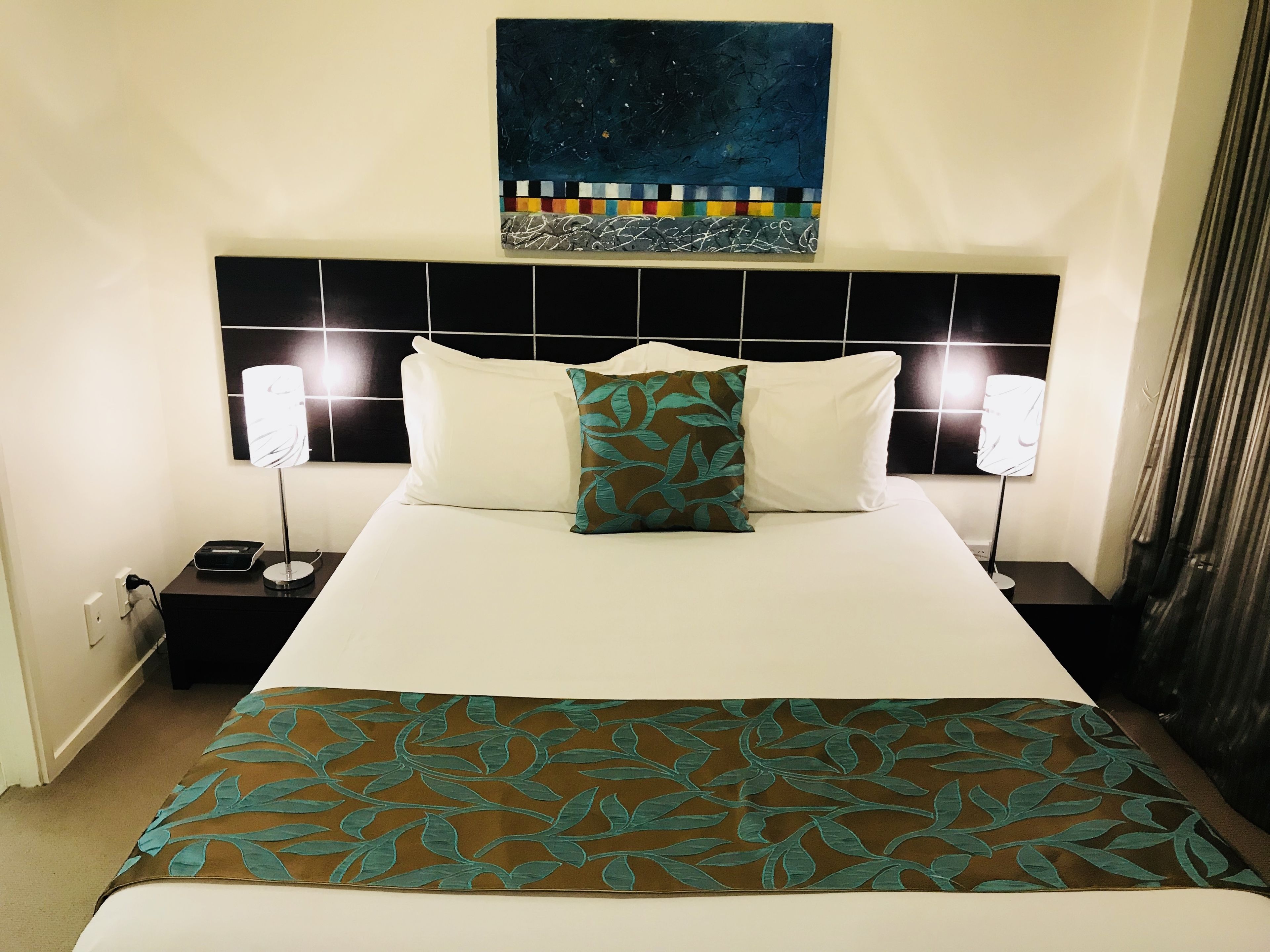 Une chambre d’hôtel avec un grand lit, deux tables de chevet, une petite table, un tableau au-dessus du lit et un rideau sur le côté droit.