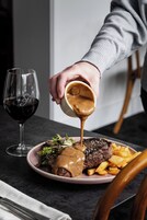 Un plato con filete cubierto de salsa, una copa de vino tinto y un tenedor sobre la mesa.