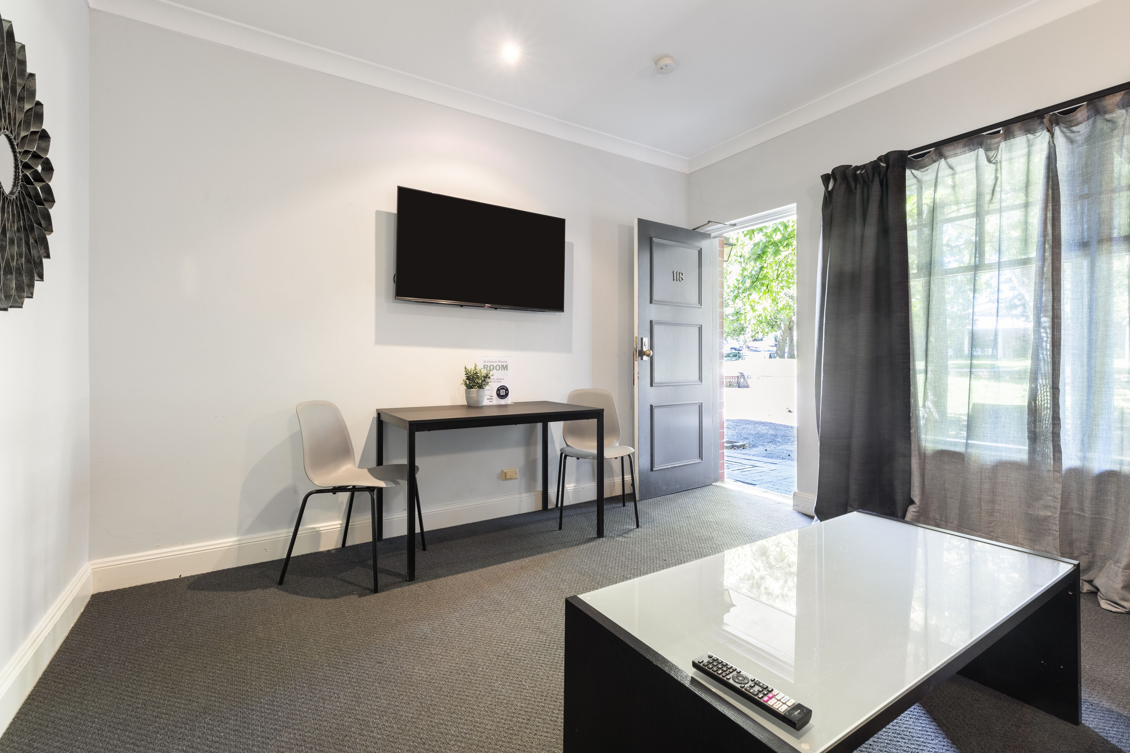 A modern hotel room with a flat-screen TV mounted on the wall, a small table with two chairs, a glass coffee table, and a door leading to an outdoor area.