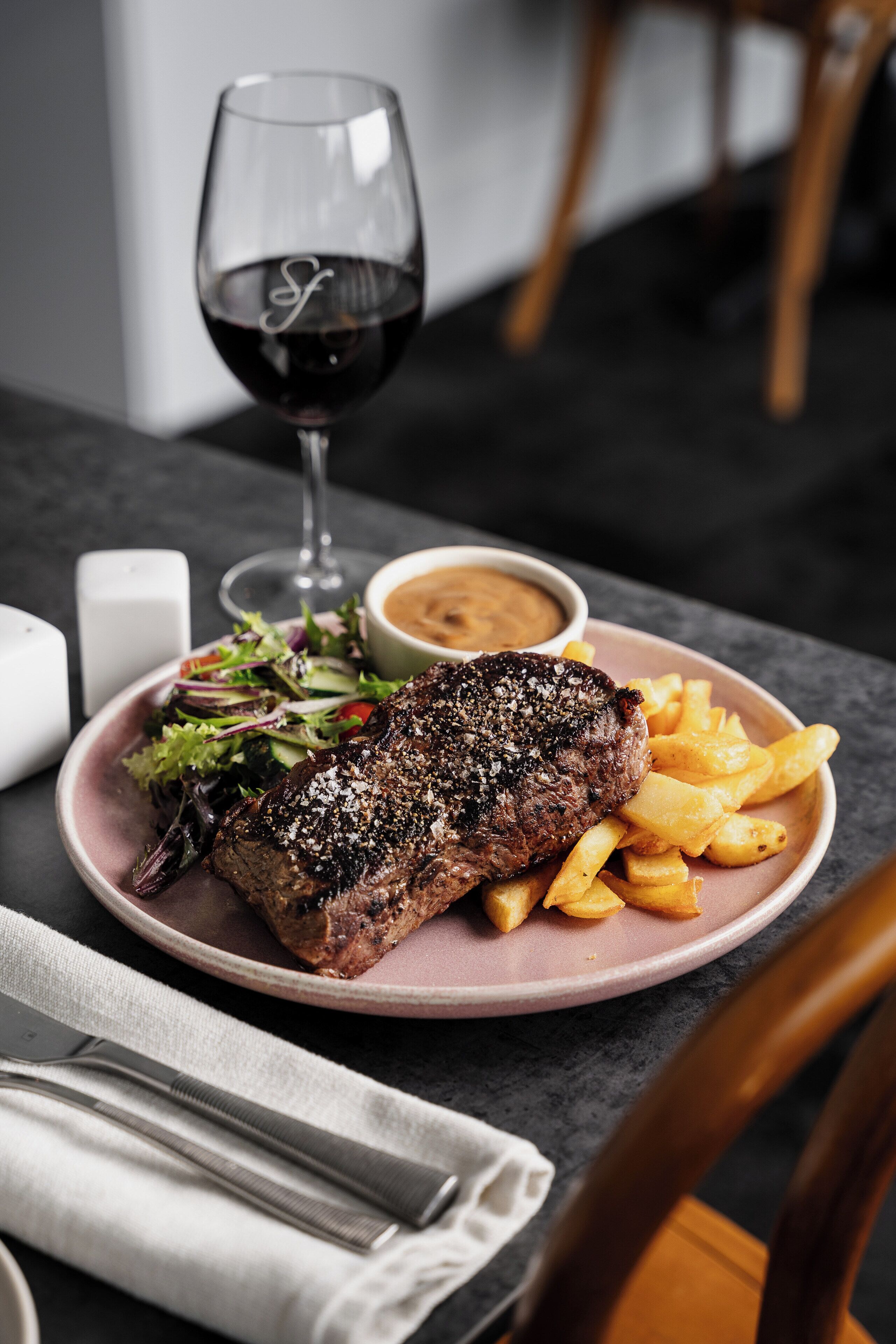 Ein Teller mit Steak, Pommes und Salat, ein Glas Rotwein und eine kleine Schale Soße auf einem Tisch.