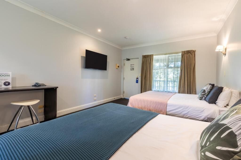 Habitación de hotel con dos camas, un escritorio con silla, televisor montado en la pared y una ventana con cortinas.
