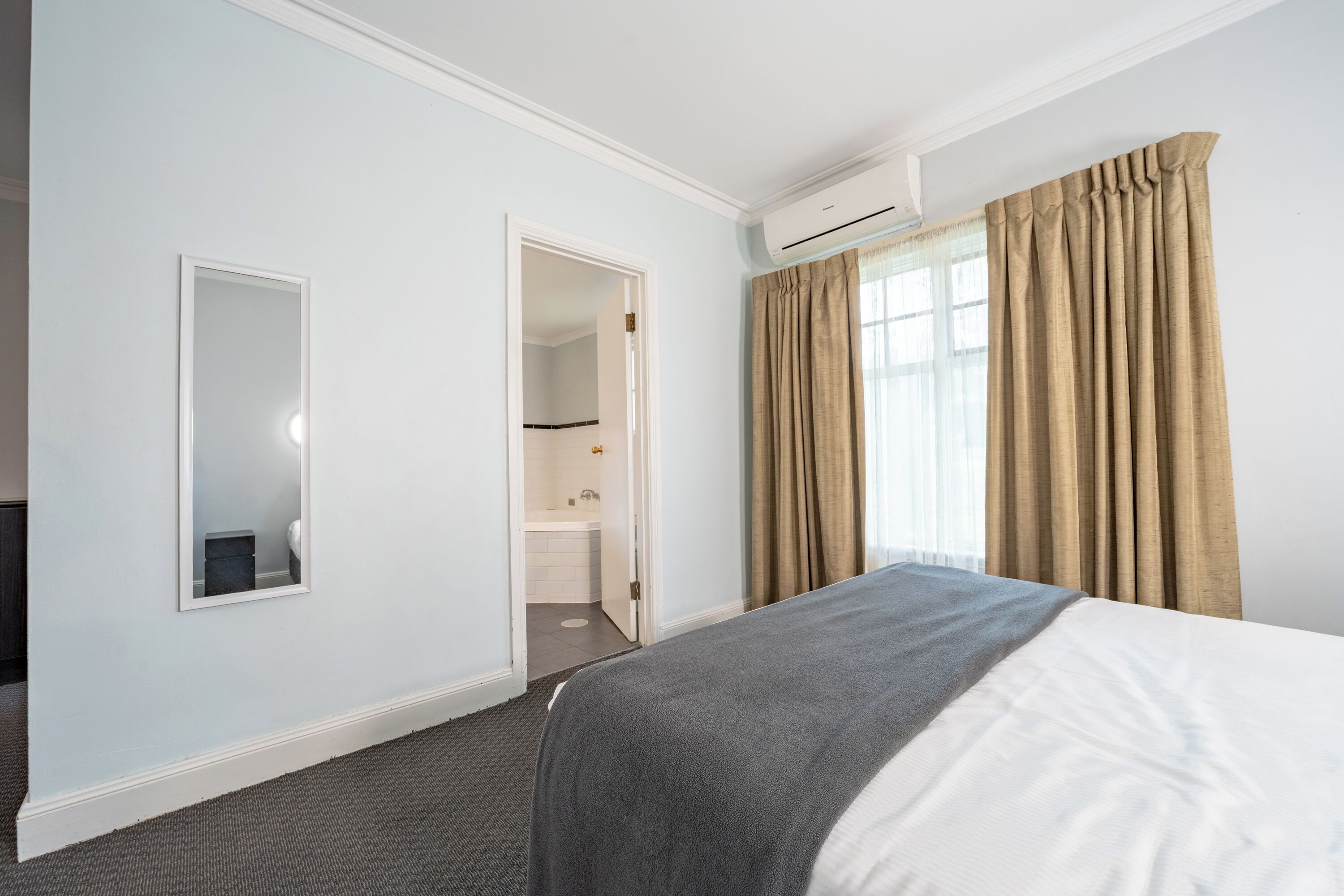 A hotel room with a bed, a mirror, a bathroom visible through an open door, and beige curtains.