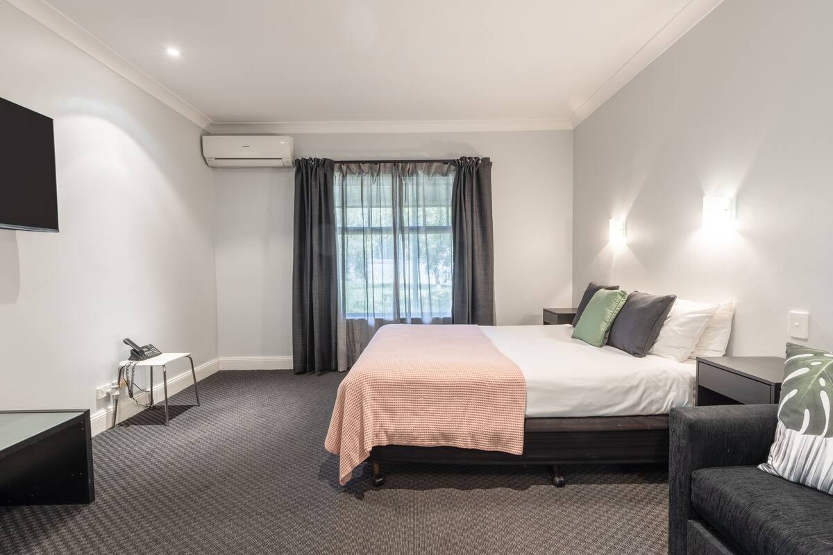 A hotel room with a bed, bedside tables, a desk, a chair, and a TV on the wall.
