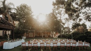 Un espace de restauration en plein air, avec des tables dressées pour un repas, entouré d’arbres et bordé d’un bâtiment à la façade en pierre.