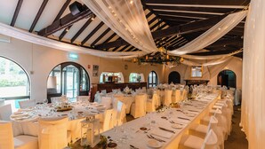 Une salle de banquet avec de longues tables préparées pour un événement formel, agrémentée de nappes et de chaises blanches, et dotée d’un plafond haut avec des poutres apparentes et des étoffes suspendues.
