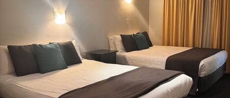 Habitación de hotel con dos camas, cada una con cabecero oscuro y ropa de cama blanca. Hay luces fijadas a la pared y una ventana con cortinas.