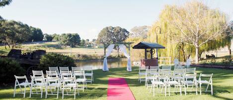 Salón de bodas al aire libre con pasillo de alfombra roja, sillas blancas, un pabellón y un estanque al fondo.