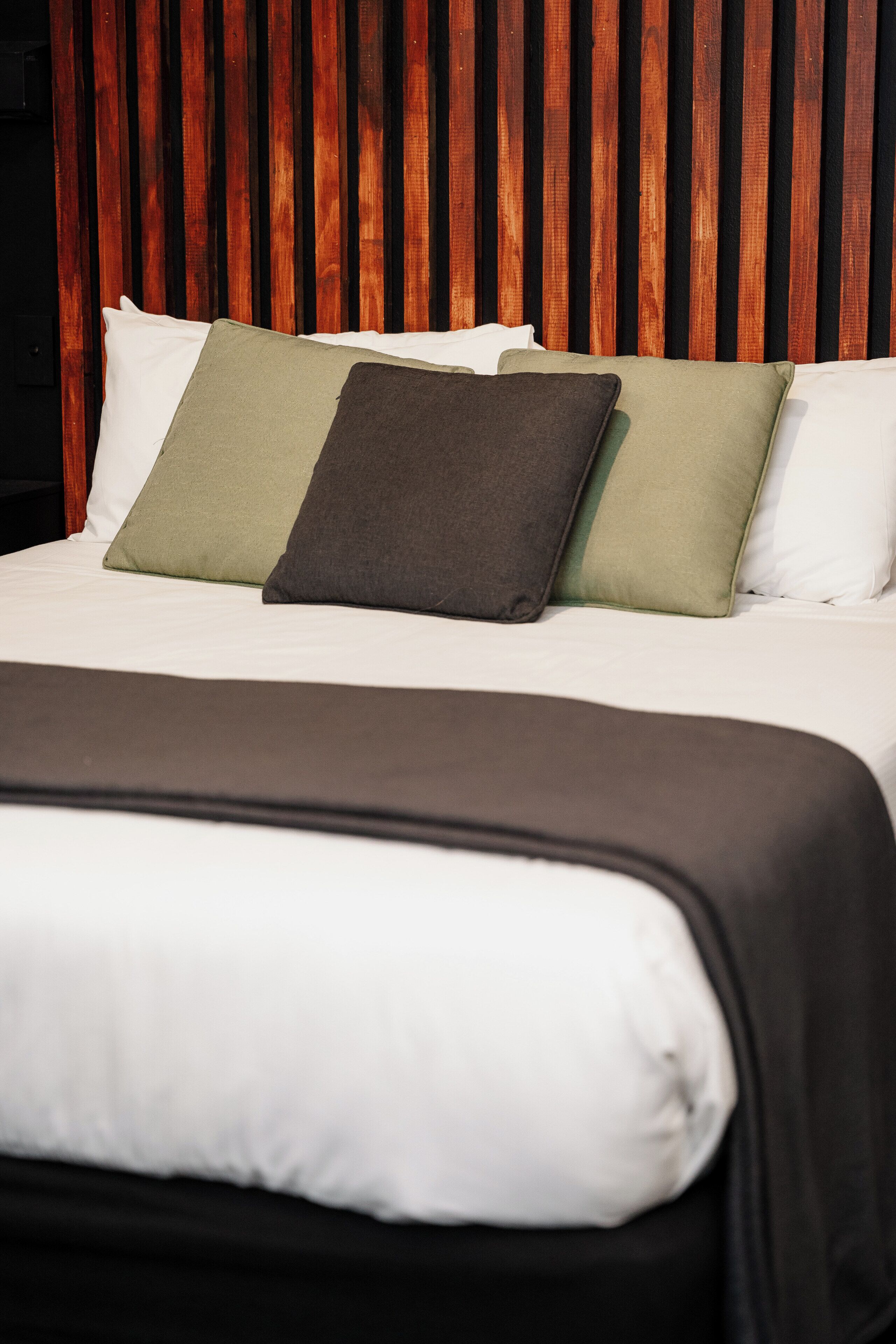 Una cama con cabecera de madera, ropa de cama blanca y tres almohadas de colores diferentes.