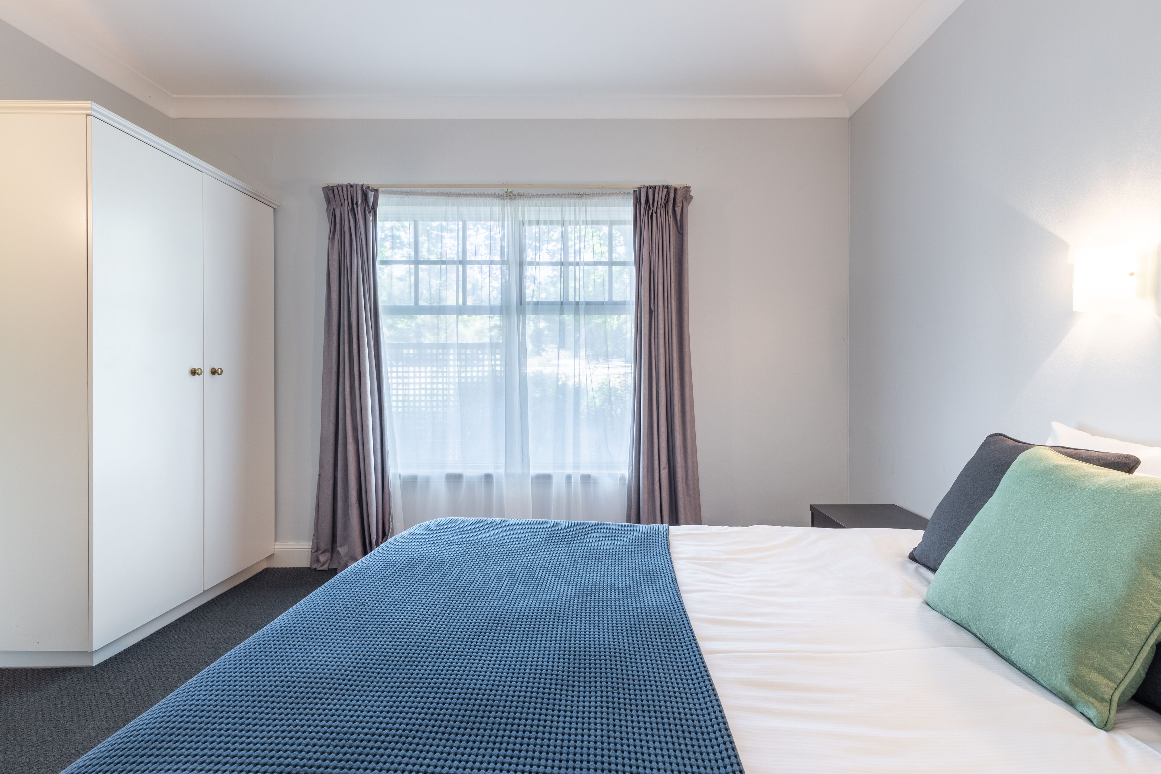 Un dormitorio con cama, armario, ventana con cortinas y mesita de noche.