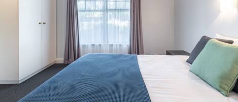 Un dormitorio con cama, armario, ventana con cortinas y mesita de noche.