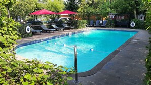 Una piscina rectangular con borde de azulejos azules, rodeada por una terraza de concreto. Hay sillas de descanso con sombrillas rojas y un edificio pequeño al fondo.