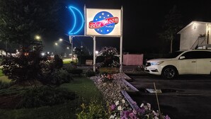 Letrero de motel con una luna creciente y estrellas, rodeado por un jardín ajardinado con flores y un automóvil blanco estacionado cerca.