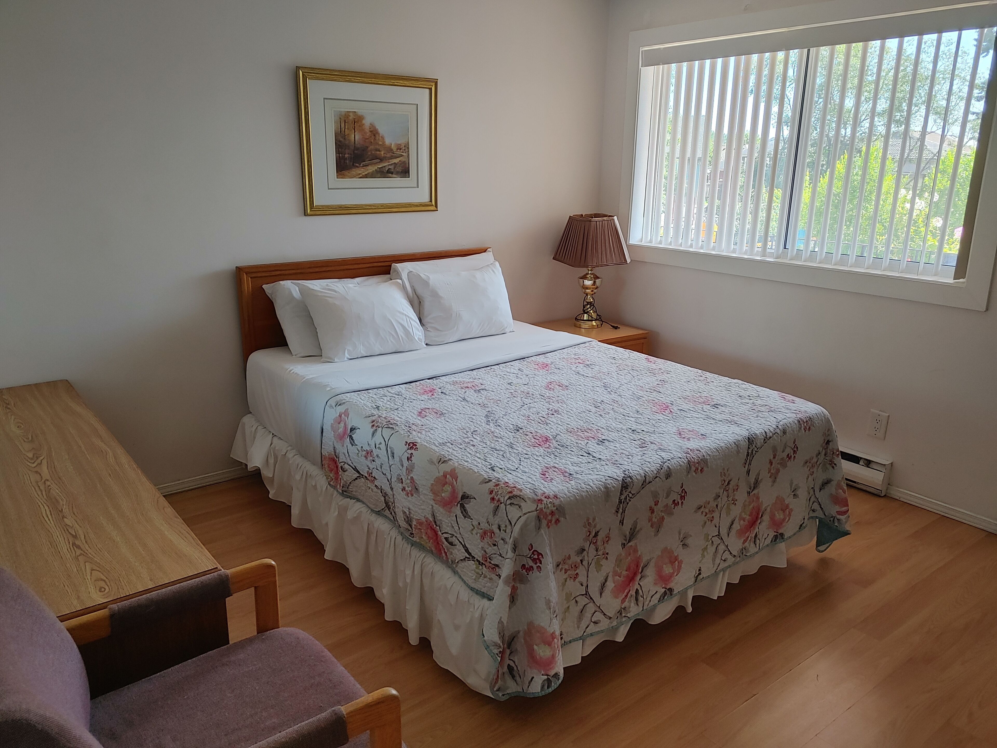 Un dormitorio ordenado con cama, mesita de noche con lámpara, una silla y ventana con persianas.