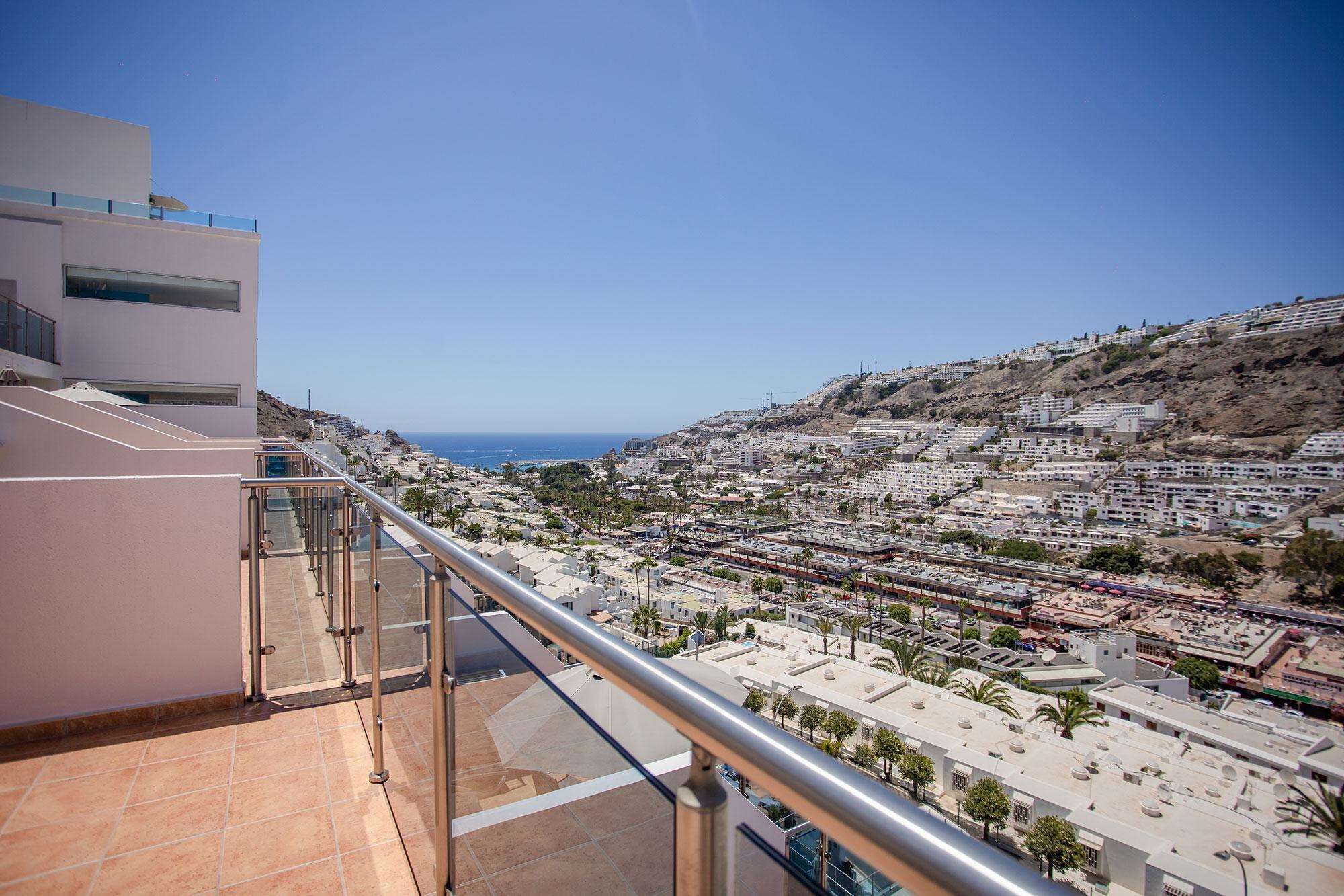 Un balcón con vistas despejadas a un pueblo costero, que muestra una mezcla de edificios residenciales y una colina destacada.