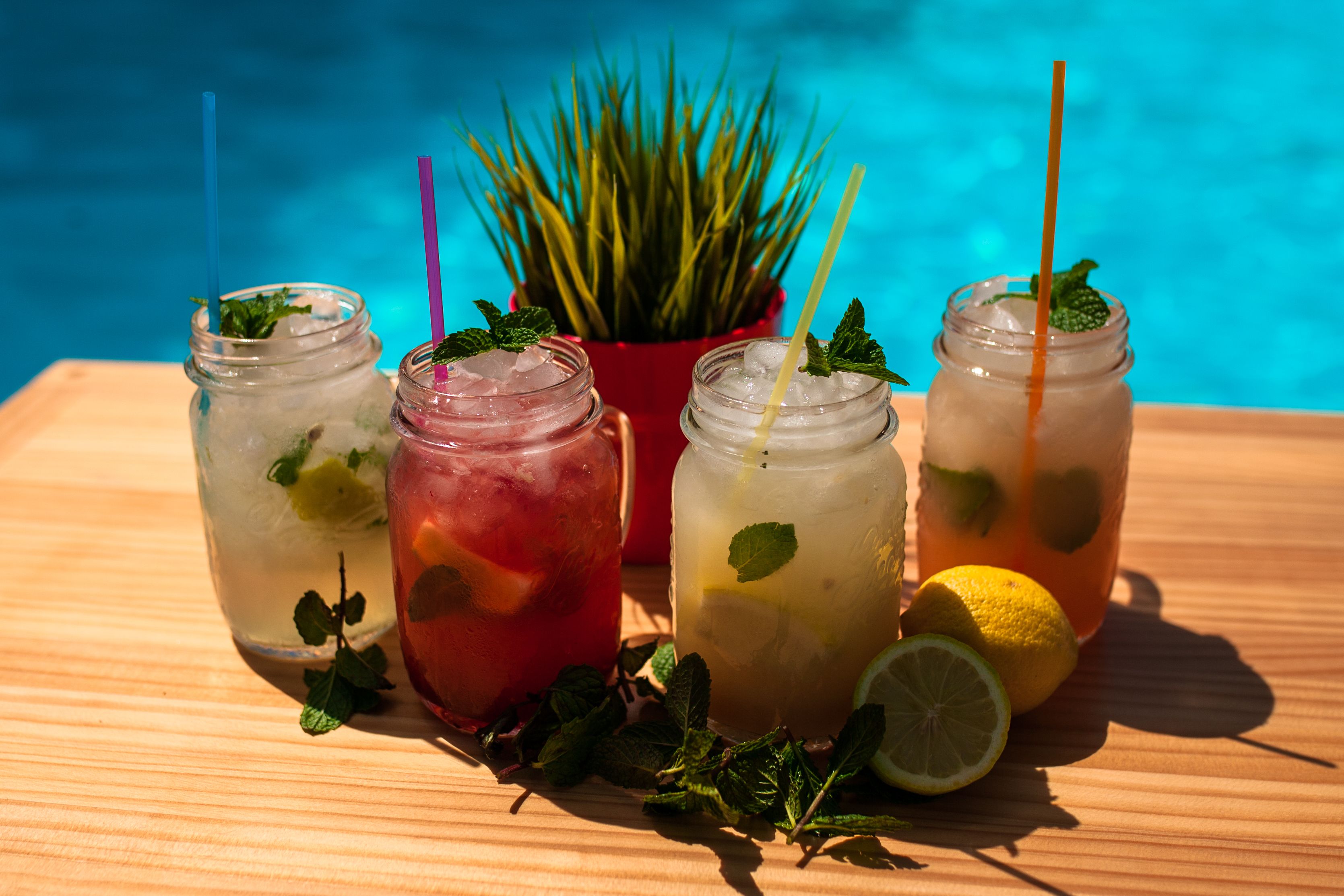 Four mason jars with drinks, mint leaves, and ice, placed on a wooden table with a pool in the background.