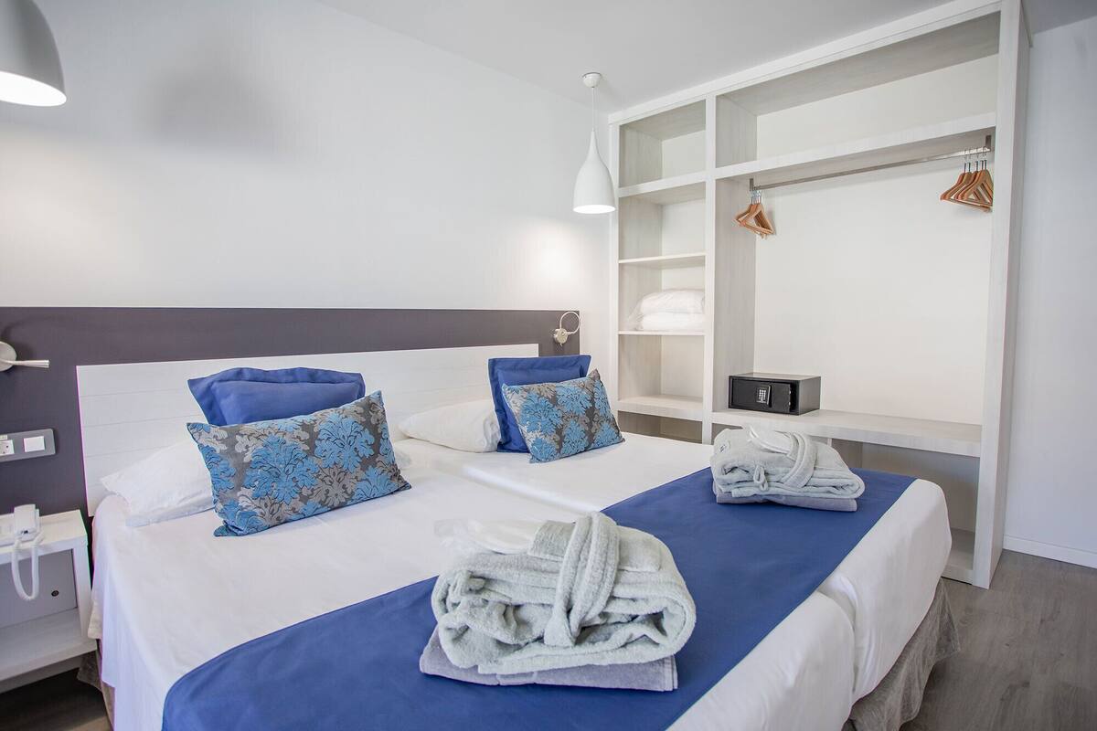 Ein Hotelzimmer mit zwei Betten, jedes mit blauen und gemusterten Kissen, einer blauen Tagesdecke und einem Handtuch auf dem Bett. Es gibt einen weißen Nachttisch mit einem Telefon und einen kleinen Tisch mit einem Radio.