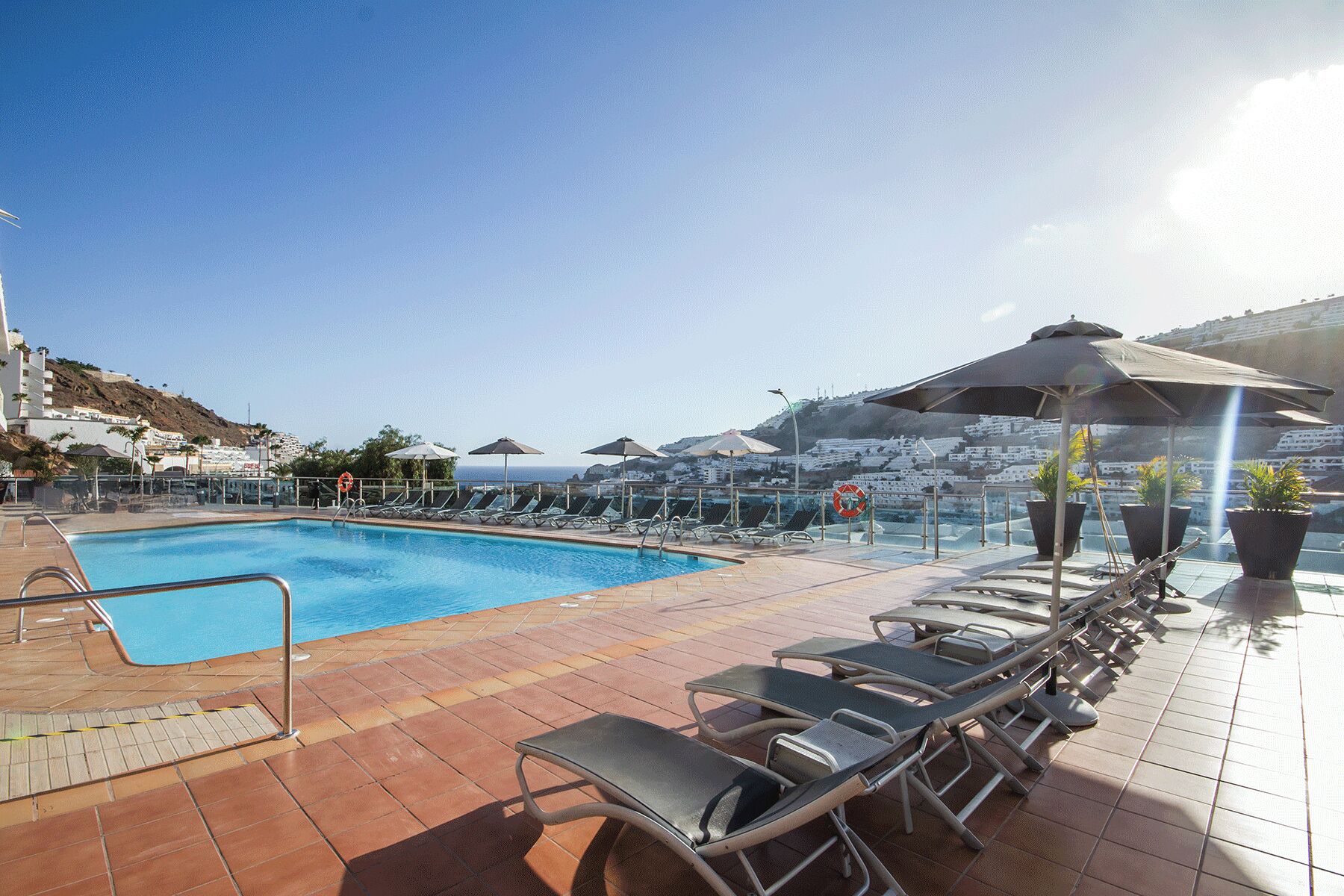 A swimming pool with sun loungers and umbrellas, surrounded by tiled flooring and a clear sky.