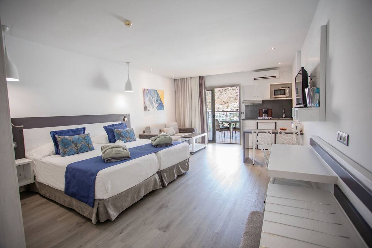 Ein modernes Hotelzimmer mit einem großen Bett, einem separaten Schreibtisch, einer Küchenzeile und einem Balkon.