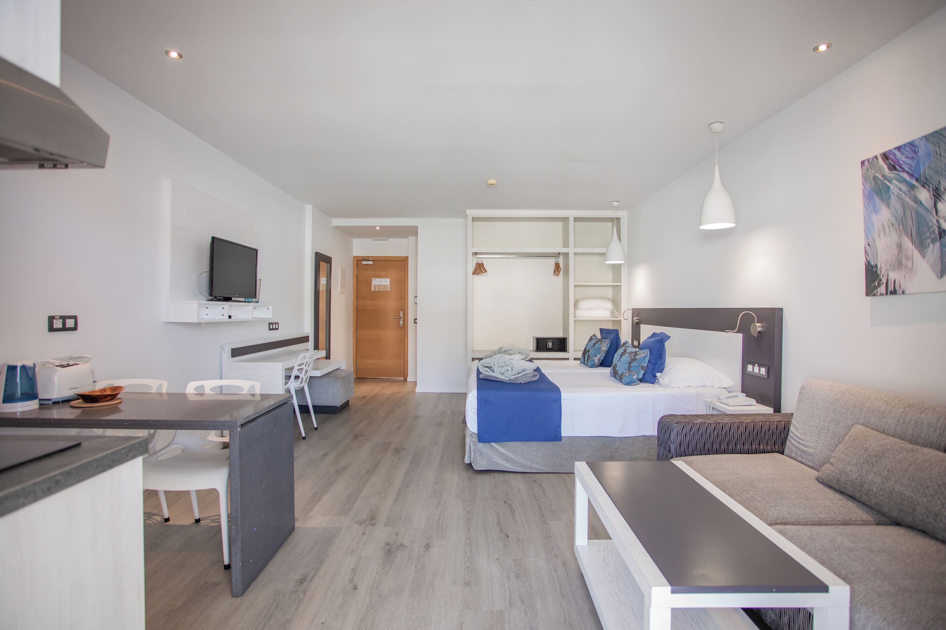 Ein modernes Hotelzimmer mit einer Küchenzeile, einem Bett mit blauer Bettwäsche, einem grauen Sofa und einem Bild an der Wand.