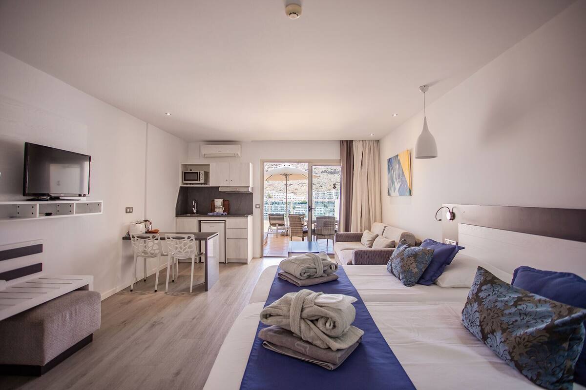 Ein Hotelzimmer mit Fernseher, einer Küchenzeile, einem Essbereich und einem Bett mit Handtüchern und Kissen.