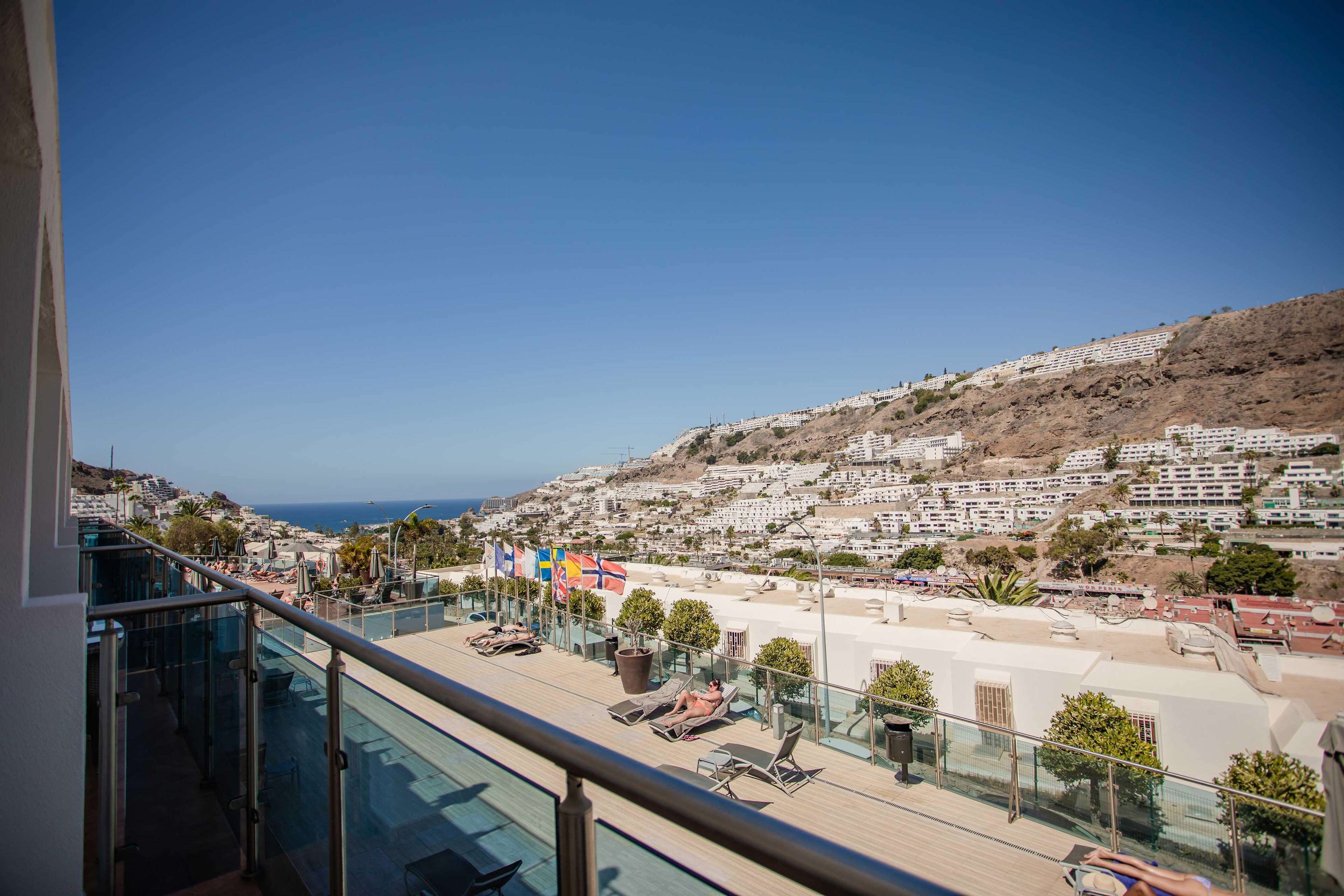 A balcony with a clear view of a coastal town, lounge chairs, and a swimming pool.