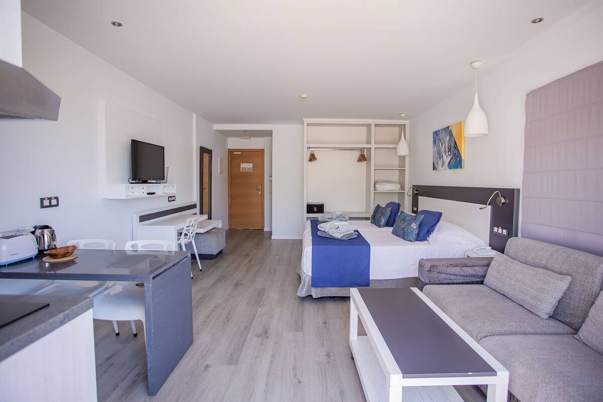 Ein modernes Hotelzimmer mit einer Küchenzeile, einem Bett mit blauer Bettwäsche, einem grauen Sofa und einem an der Wand befestigten Fernseher.