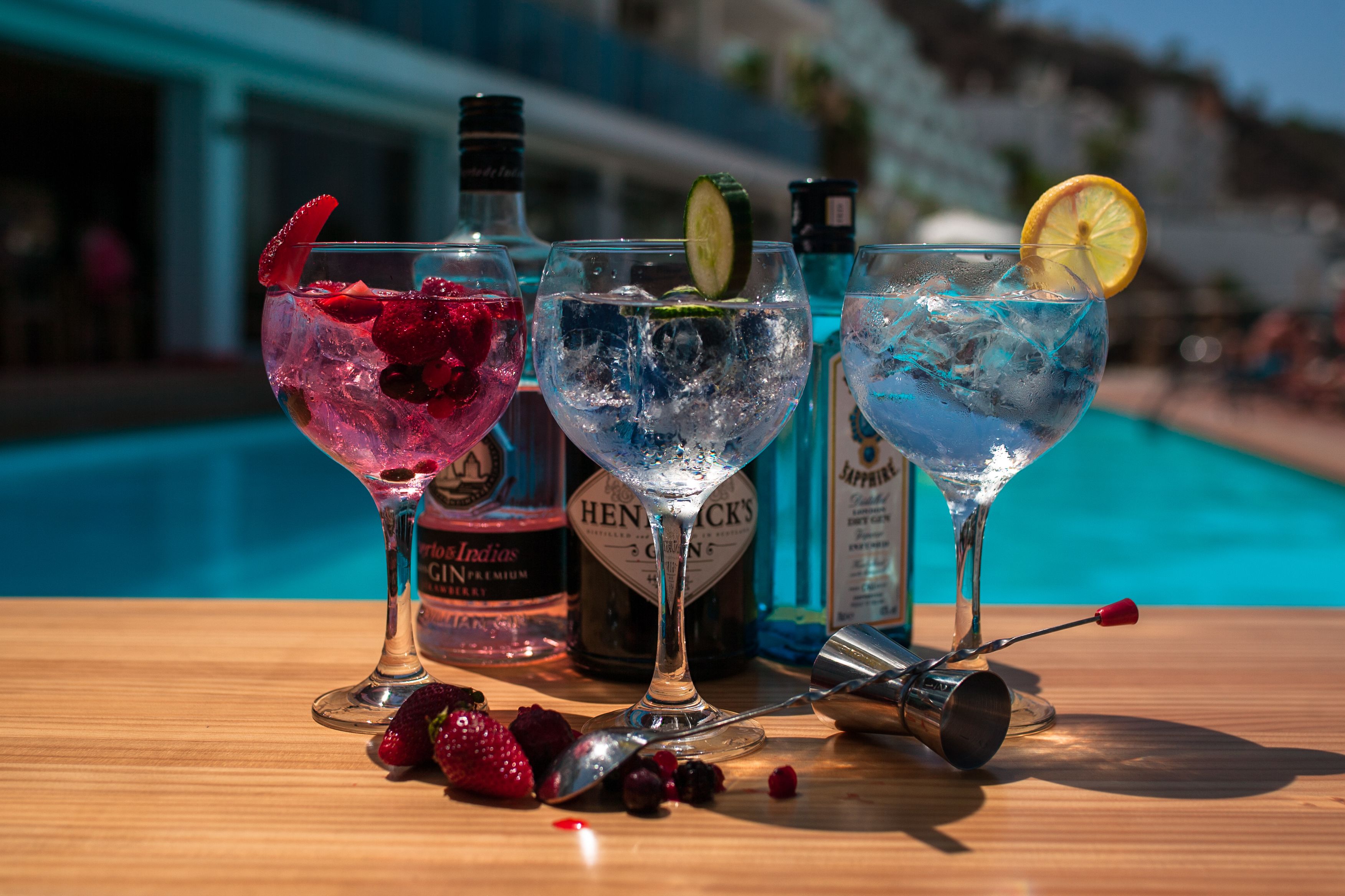 Three cocktails on a wooden table with ice and garnishes, a bottle of Hendrick's Gin, and a cocktail shaker.