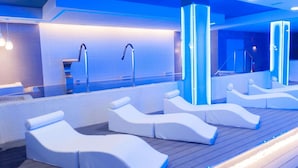 Ein moderner Wellnessbereich mit mehreren weißen Liegen, einem großen Spiegel und einer blau beleuchteten Wand.