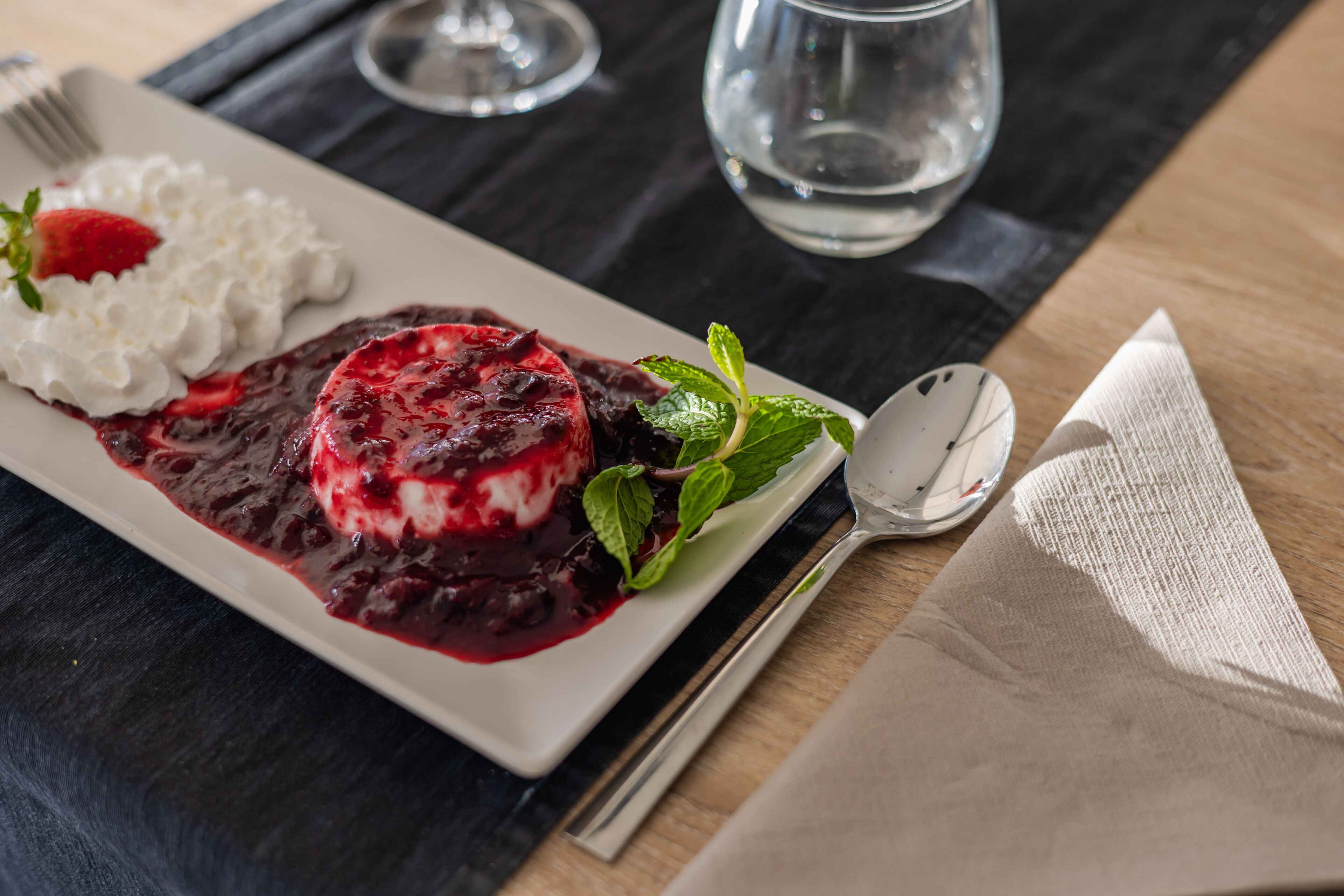 Un plato de postre con compota de frutos rojos y crema batida, acompañado de una ramita de menta, sobre una mesa oscura con un vaso de agua y cubiertos.