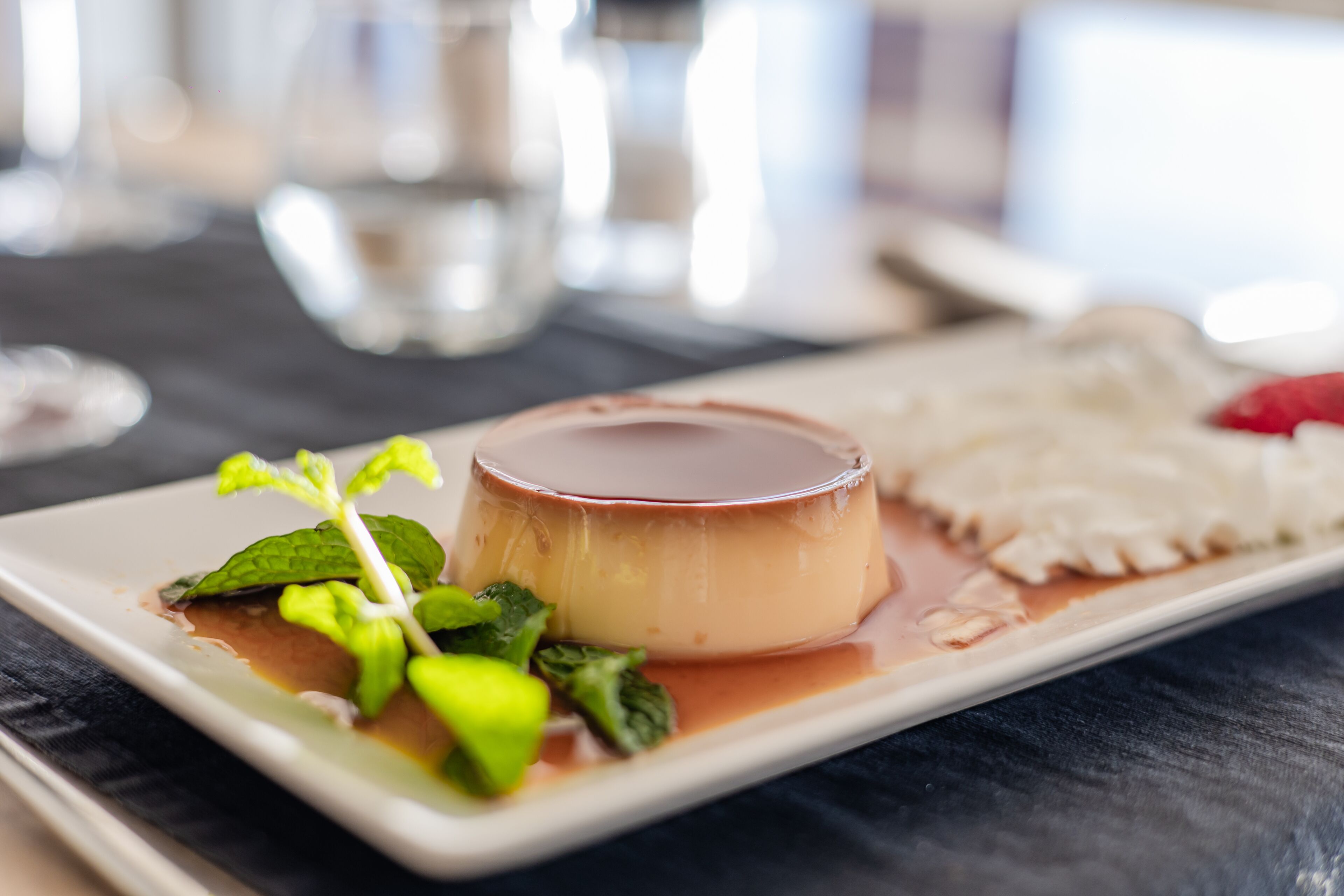 Un plato de postre con un flan de caramelo, hojas de menta y una rebanada de pastel sobre un plato blanco rectangular.