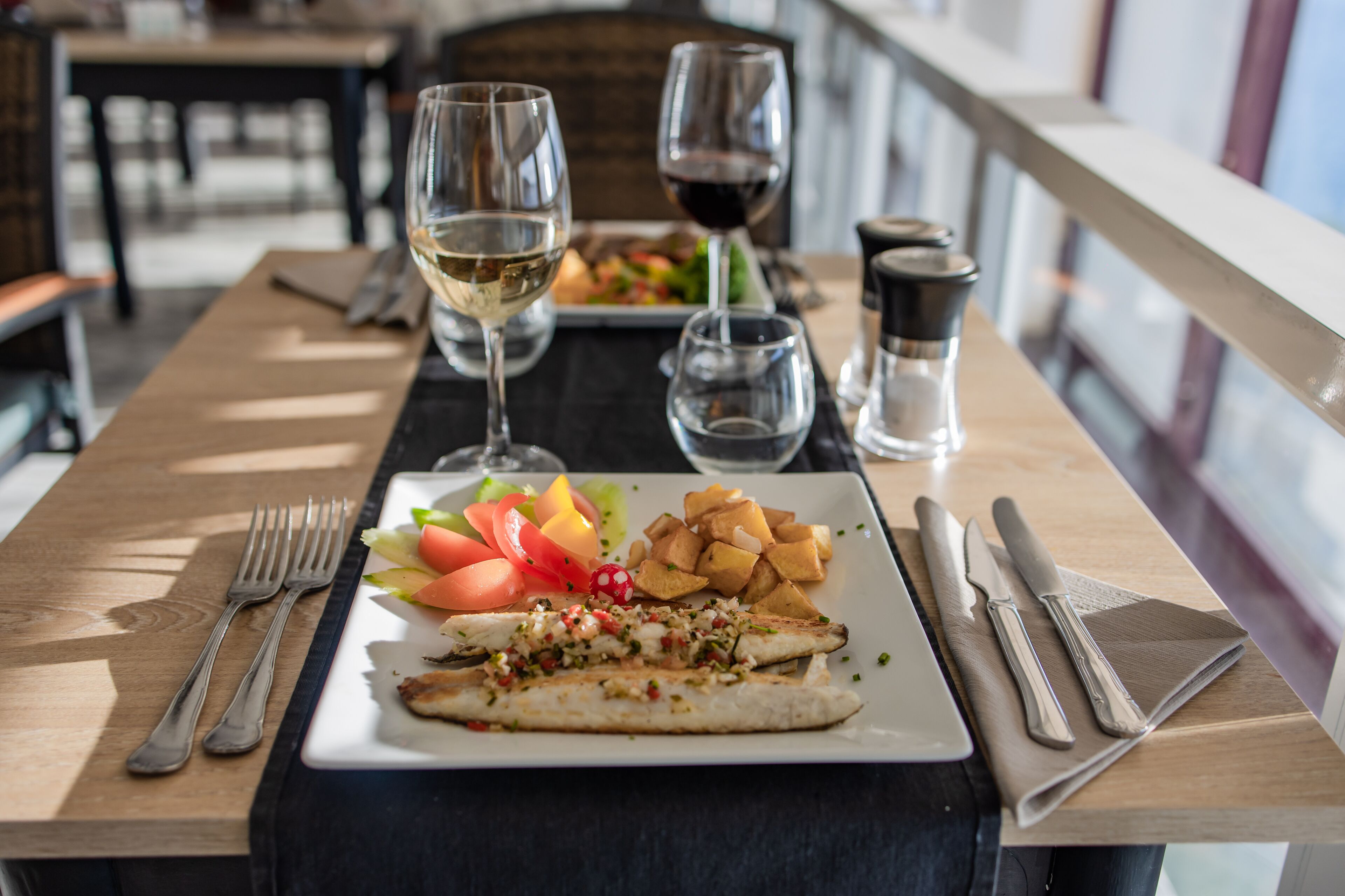 A dining table with a plate of food, including a fish fillet, vegetables, and chips, along with wine glasses and cutlery.