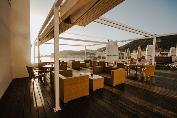 A covered outdoor seating area with wicker furniture and umbrellas, overlooking a body of water and hills.