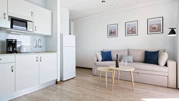 Eine moderne Küche mit weißen Schränken, einer Mikrowelle, einer Kaffeemaschine und einem Kühlschrank. Es gibt ein beiges Sofa mit blauen Kissen sowie einen kleinen runden Tisch mit zwei Gläsern und einer Flasche.