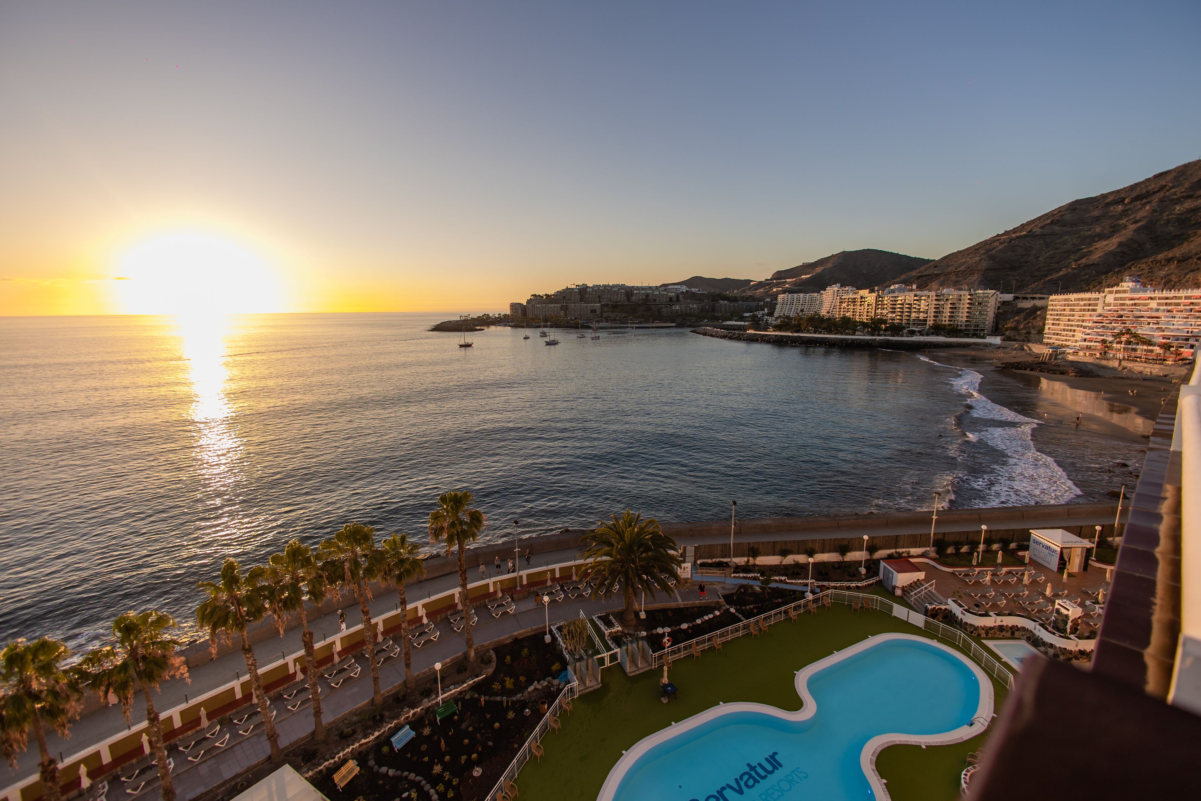 A resort with a swimming pool, beach access, and a view of the sunset over the ocean.
