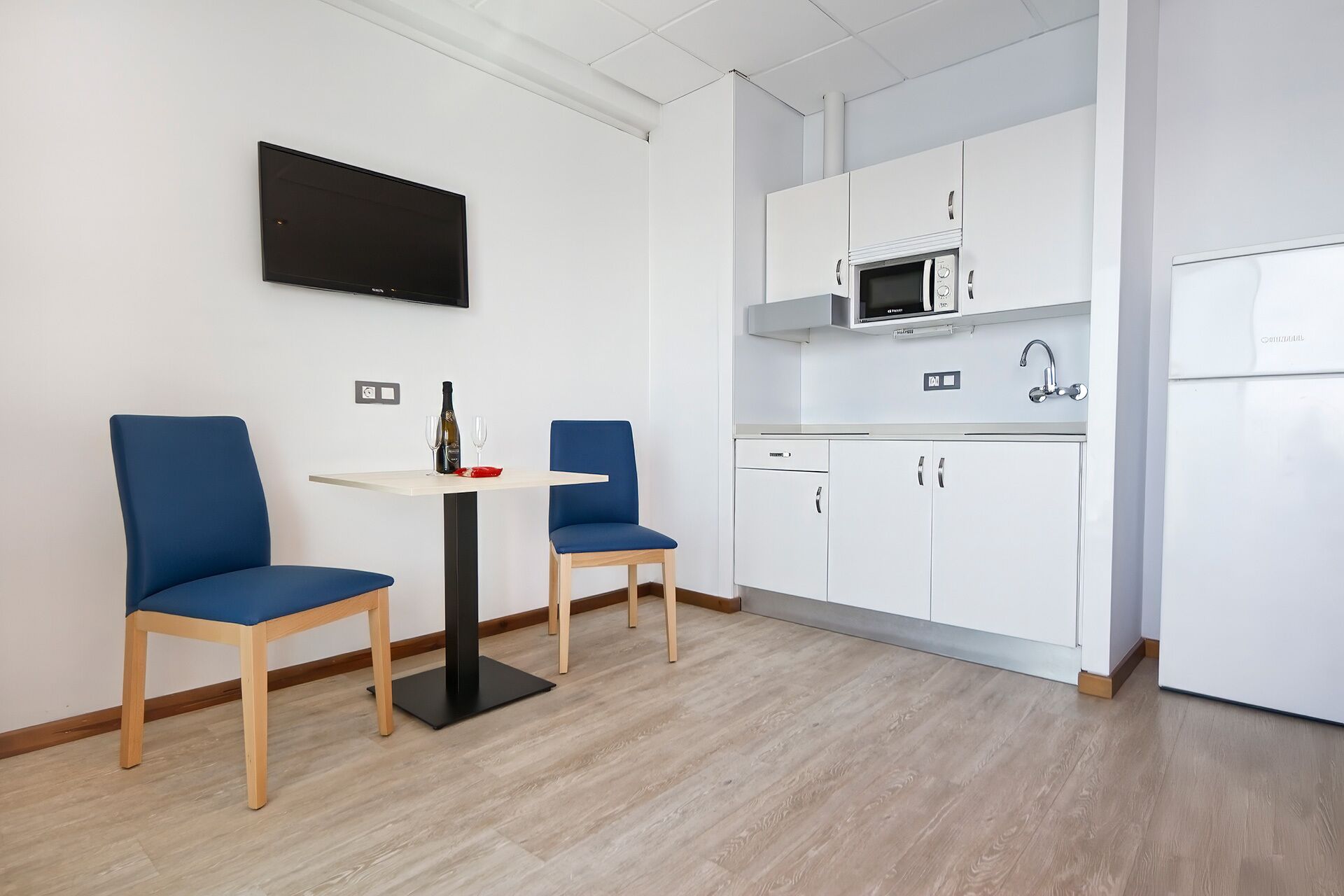 A modern kitchen with white cabinets, a microwave, and a refrigerator. There is a small dining area with two blue chairs and a table.