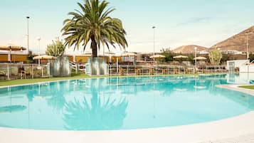 Una piscina amplia y cristalina con una palmera y sombrillas al fondo.
