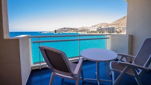 A balcony with two chairs and a small table, overlooking a turquoise sea and a coastal town. - Servatur Green Beach (Mogan)