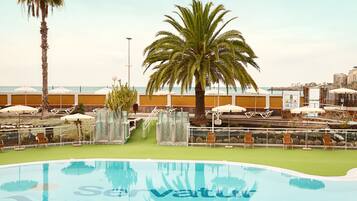 Zona de piscina con sillones de descanso y sombrillas, una palmera y vista al mar.